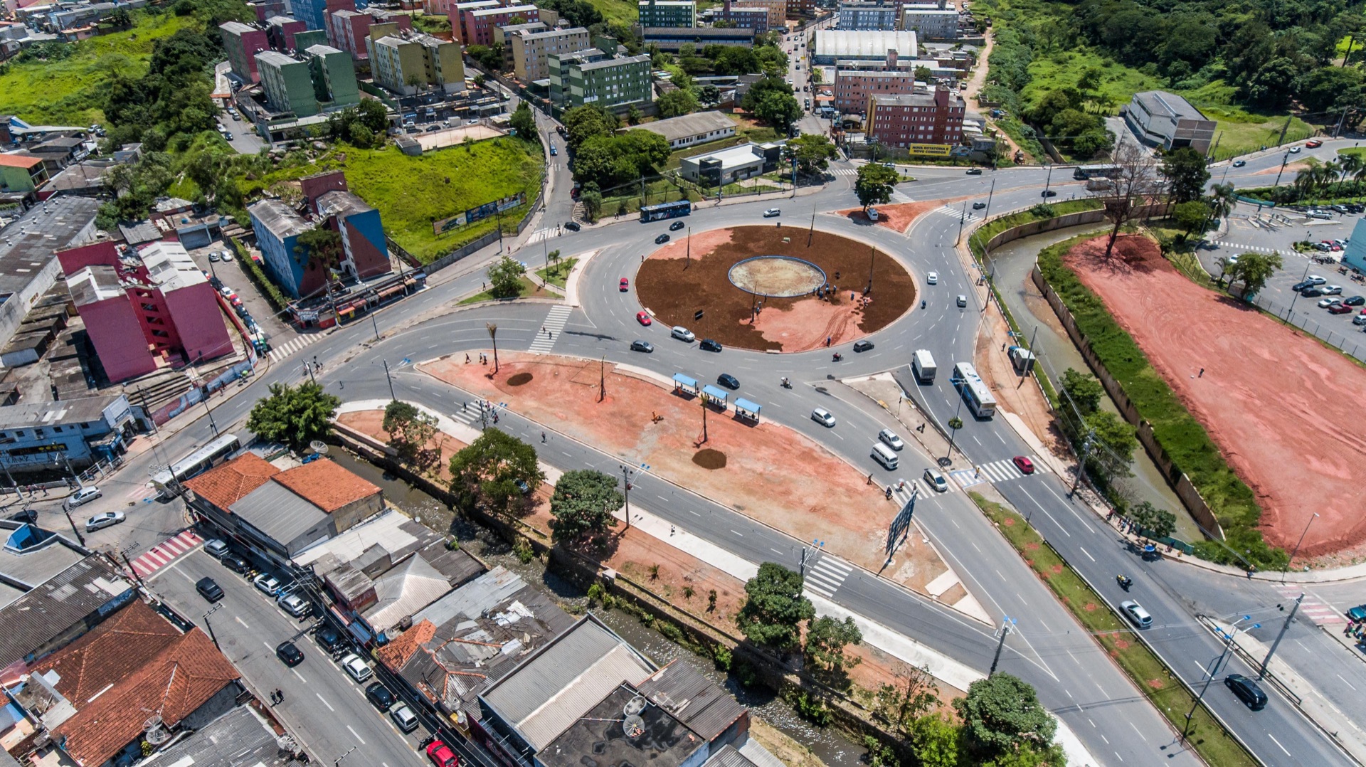Vista aérea de uma rotatória e de uma obra viária em andamento, com ruas e edifícios ao redor